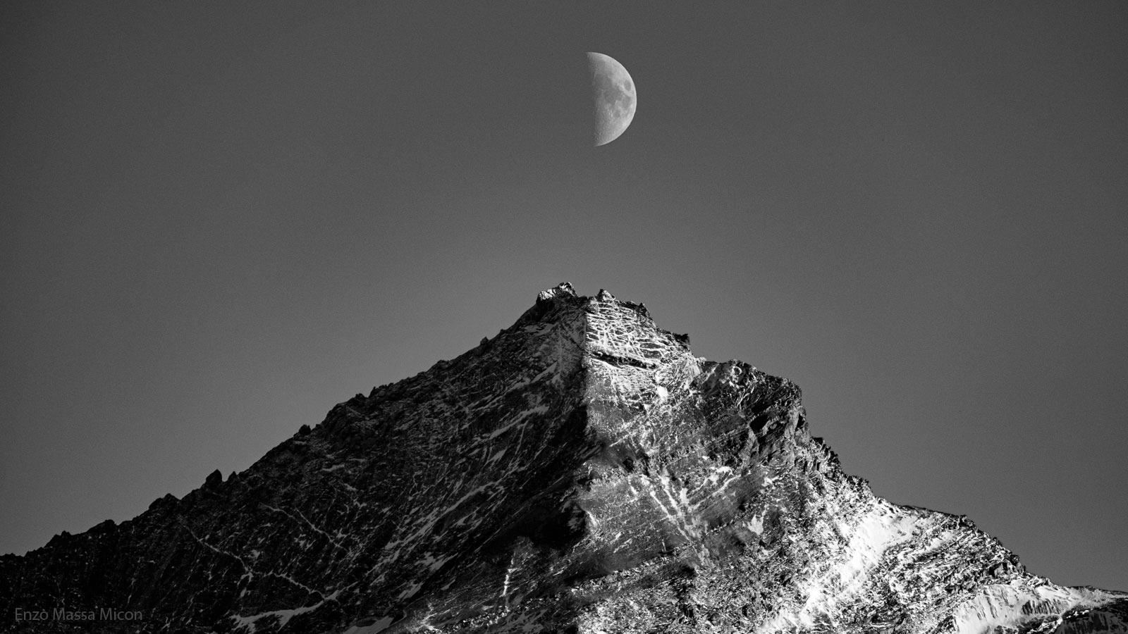 صورة بيضاء وسوداء يظهر فيها القمر وجبل. كلاهما نصف مُضائَين بواسطة الشمس، مع كون النصف الآخر مُظلّلاً. يكون نصف القمر فوق قمّة الجبل مُباشرةً.