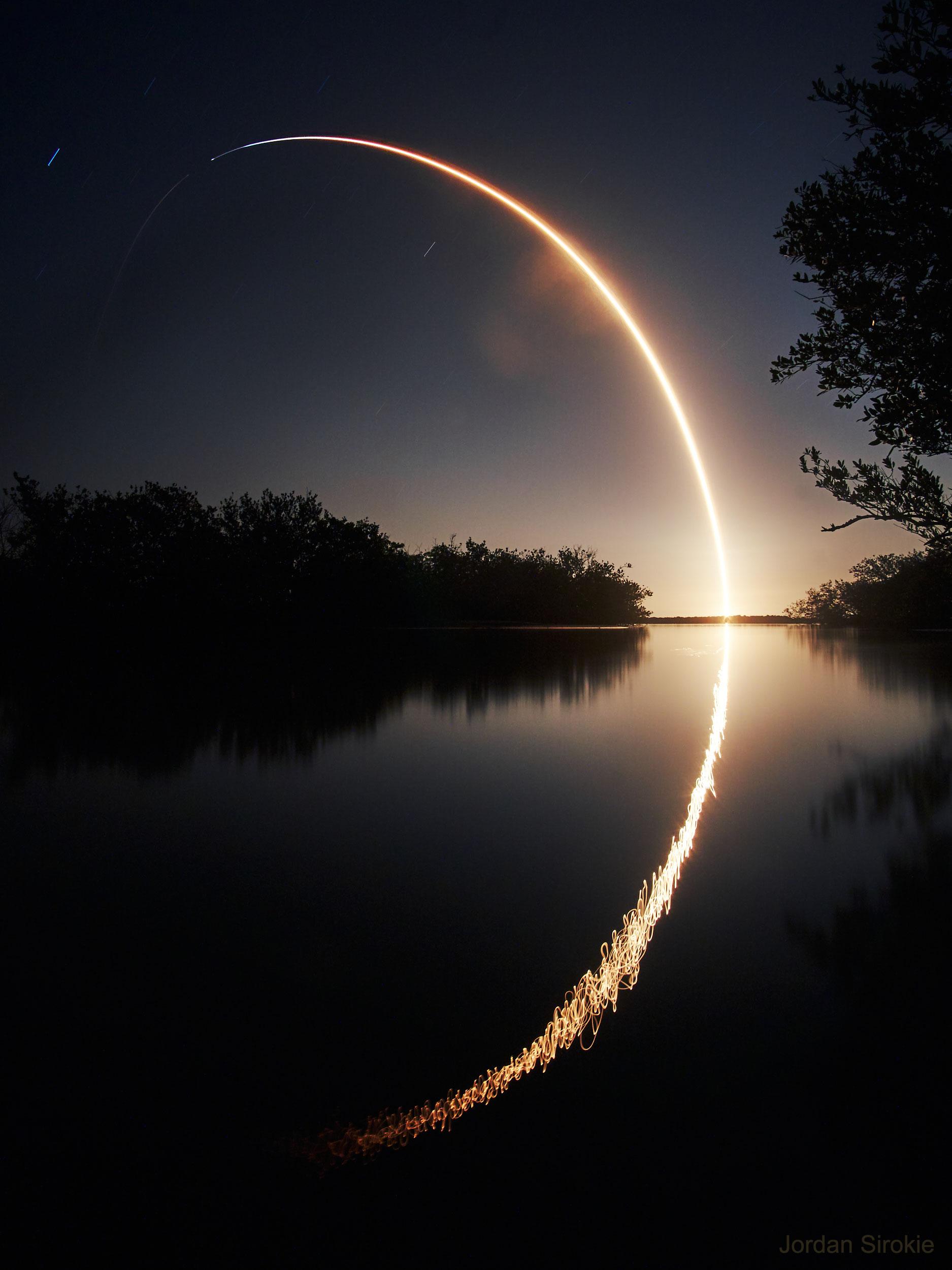 صورة طويلة التعريض تظهر مسار صاروخ منطلق من سطح الأرض راسماً فوس في السماء بينما ينعكس ذلك على صفحة مسطّح مائي
