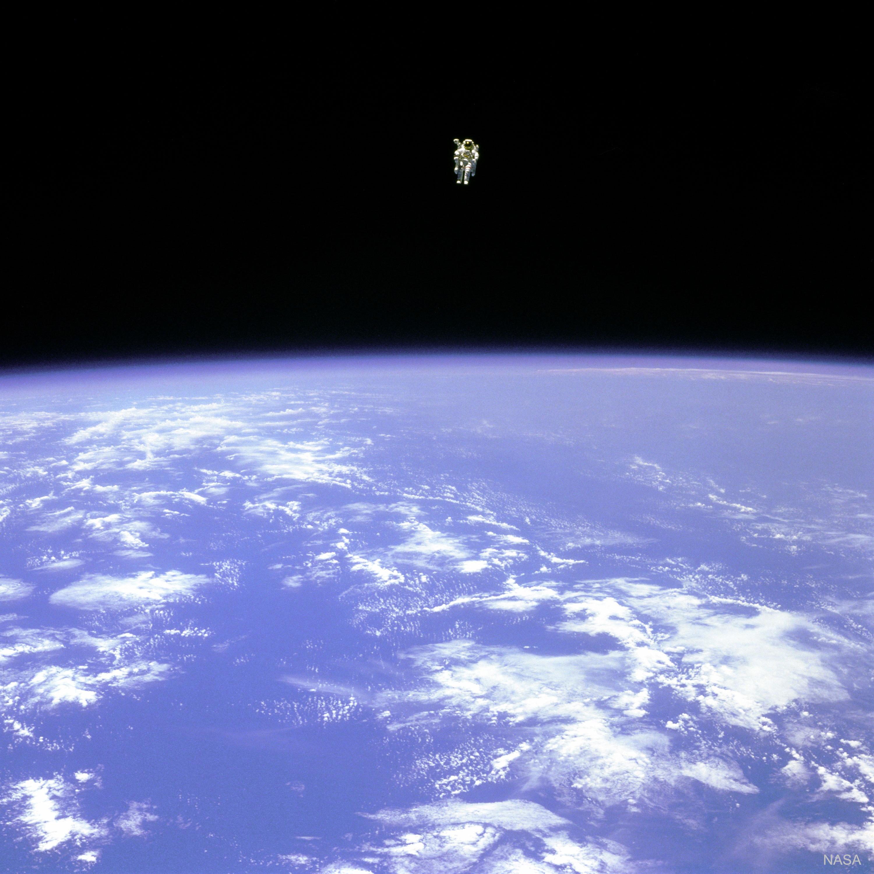 رائد فضاءٍ يحوم فوق الأرض. في الجزء العلوي من الصورة، يُرى رائد الفضاء قبالة عتمة الفضاء. في الجزء السفلي من الصورة، تكون الأرض زرقاء ساطعة مع سُحُبٍ بيضاء.