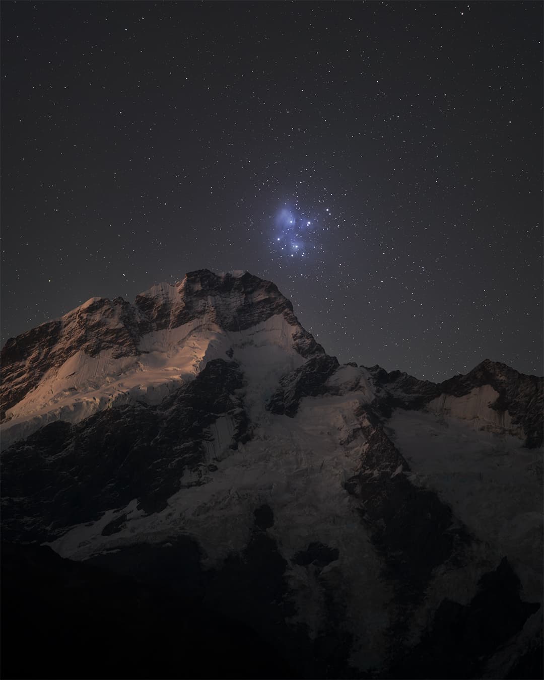 تجمّع نجمي تغمره سدم مزرقّة ضمن سماءٍ مرصّعة بالنجوم تعلو حافّة جبليّة تكسوها الثلوج