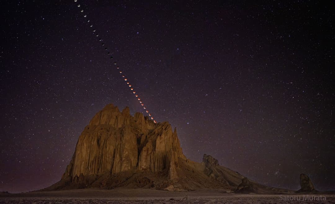 مشهد صحراويّ مع معلم أرضيّ بركانيّ كبير يبرز في المنتصف. في الأعلى توجد سلسلة من أطوار الخسوف تنتقل من أعلى اليسار نزولاً باتّجاه قمّة المعلم البركانيّ المركزيّ. تُظهر الأطوار انتقال القمر من بدرٍ أبيض إلى قمرٍ مظلم ومن ثمّ إلى قمرٍ أحمر بمجرّد اتّصاله بالمعلم في الصورة.